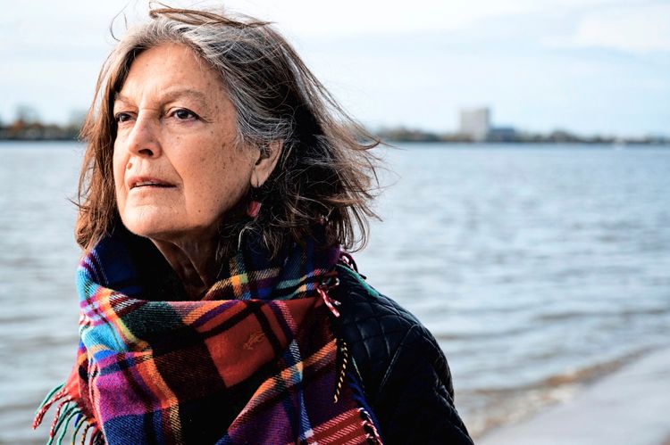 Eine ältere Frau mit grauen Haaren steht am Ufer eines Gewässers. Sie trägt einen bunten Karoschal und blickt gedankenverloren in die Ferne. Im Hintergrund ist Wasser und eine verschwommene Stadtlandschaft zu sehen.