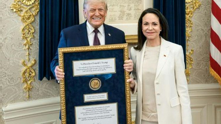 Zwei Personen stehen in einem repräsentativen Raum vor dunklen Vorhängen und halten gemeinsam einen großen gerahmten Schaukasten. Im Schaukasten befindet sich eine goldfarbene Medaille sowie mehrere Texttafeln. Rechts im Hintergrund ist ein Teil einer rot-weiß gestreiften Flagge zu sehen. Der Rahmen des Schaukastens ist goldfarben und reich verziert.