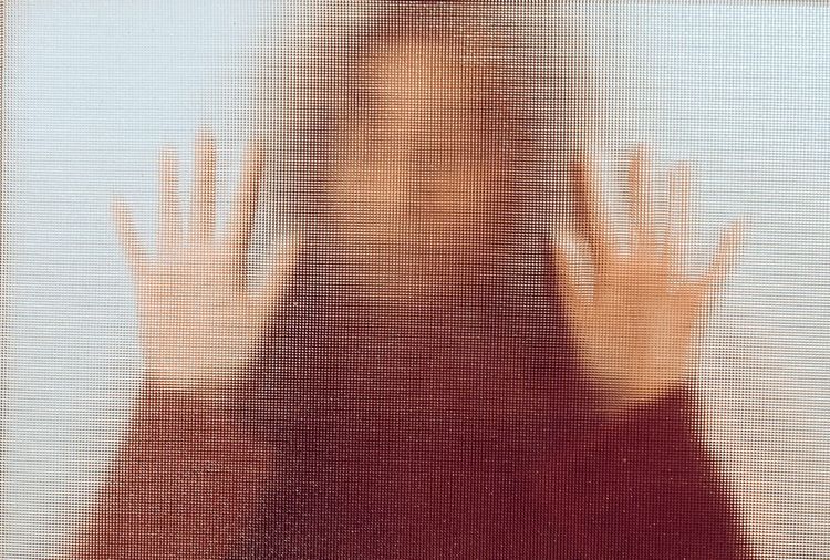 Von hinten beleuchtetes Bild der Silhouette einer Frau, die ihre Hände gegen ein Glasfenster drückt.