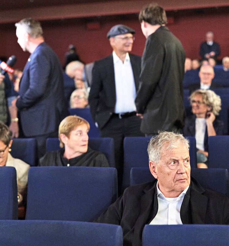 Eine Gruppe von Personen in einem Kinosaal während einer Veranstaltung. Im Vordergrund sitzt ein älterer Mann, während im Hintergrund weitere Personen stehen und sich unterhalten. Der Raum ist mit blauen Kinosesseln ausgestattet.