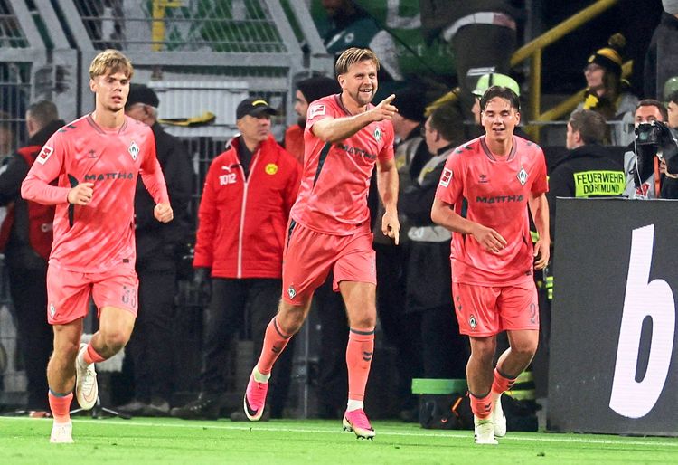Drei Fußballspieler von Werder Bremen in rosa Auswärtstrikots feiern ein Tor während eines Spiels der Bundesliga im Signal Iduna Park in Dortmund. Zuschauer und Sicherheitskräfte sind im Hintergrund zu sehen.