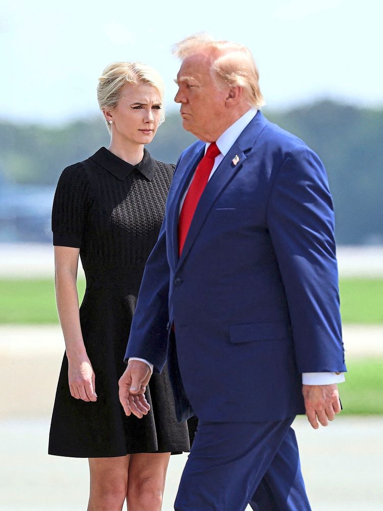 Zwei Personen sind abgebildet: Im Vordergrund ein Mann in einem blauen Anzug mit roter Krawatte, der zur Seite geht, bei dem es sich um US-Präsidenten Donald Trump handelt und im Hintergrund eine Frau in einem schwarzen Kleid, bei der es sich um seine persönliche Assistentin Natalie Harp handelt. Der Hintergrund zeigt eine unscharfe, outdoor Umgebung mit viel Himmel.