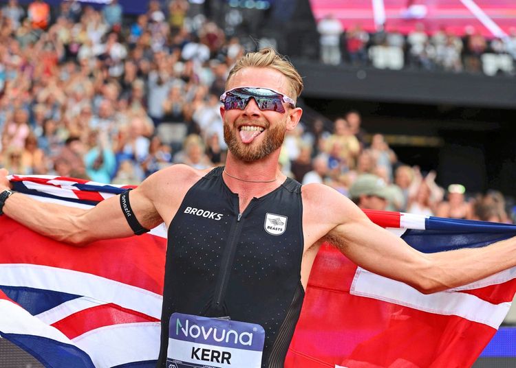 Ein Athlet in einem schwarzen Laufanzug feiert, während er die britische Union Jack-Flagge hält. Er trägt eine Sonnenbrille und zeigt seine Zunge, umgeben von jubelnden Zuschauern in einem Stadion.