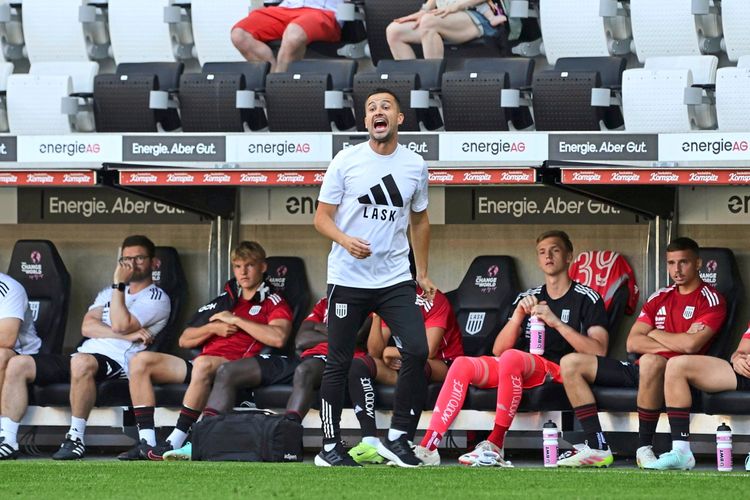 Ein Trainer des LASK steht in der Coaching-Zone vor der Ersatzbank in der Raiffeisen Arena in Linz. Auf der Bank sitzen mehrere Spieler in Trainingskleidung, im Hintergrund sind Zuschauer auf der Tribüne zu sehen.