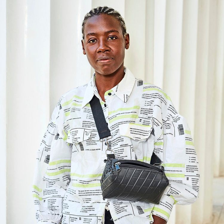 Eine Person steht vor einer weißen Säulenwand. Sie trägt ein Hemd mit grafischem Muster in Weiß, Schwarz und Gelb, eine schwarze Umhängetasche und dunkle Hose. Die Haare sind in Cornrows geflochten.