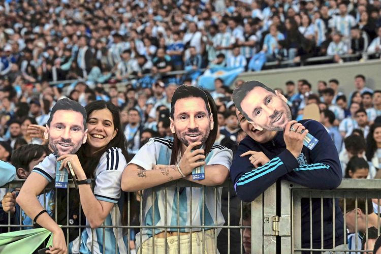 Argentinische Fans in Trikots der Nationalmannschaft halten Masken mit dem Gesicht eines bekannten Spielers, während sie vor Beginn eines Fußballfreundschaftsspiels im Stadion hinter einer Absperrung stehen.Im Hintergrund ist eine Menschenmenge zu sehen.