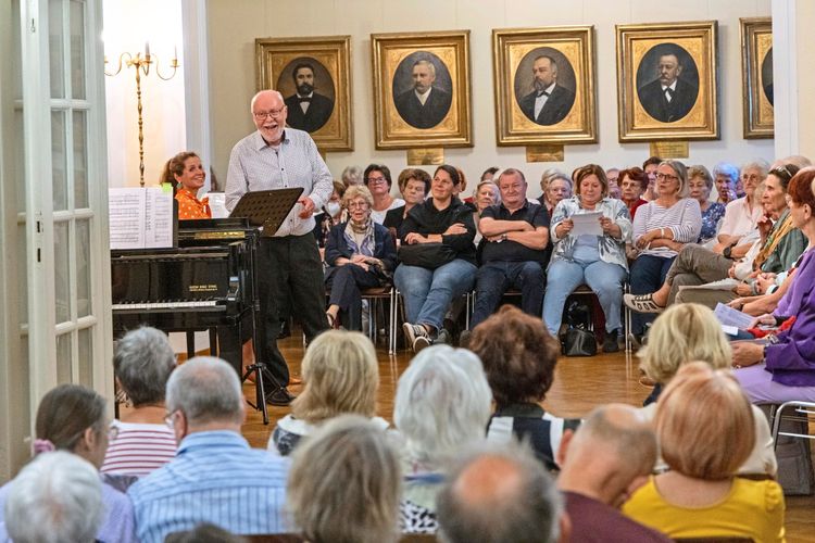 Eine kulturelle Veranstaltung in einem Saal mit zahlreichen Zuschauern. Ein älterer Mann steht lächelnd vor einem Klavier, daneben eine Frau in orangefarbener Bluse. Im Hintergrund sind gerahmte Porträts an der Wand zu sehen.