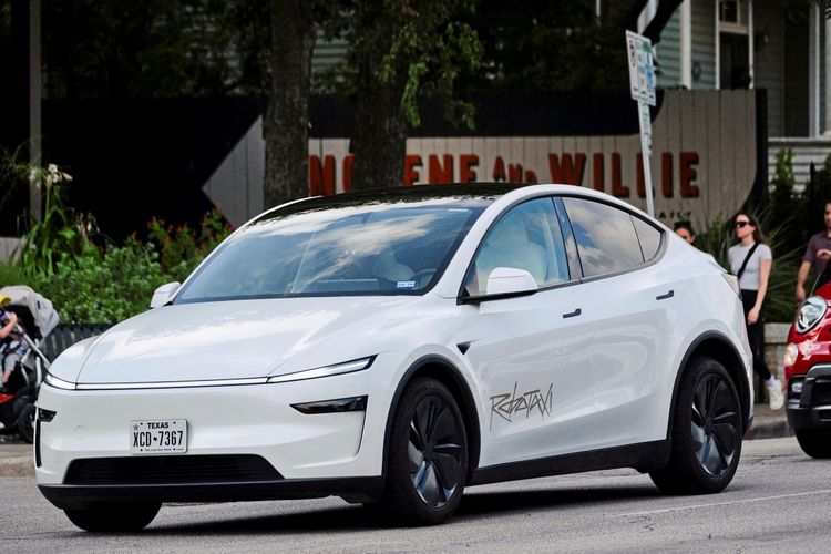 Ein weißes Tesla-Robotaxi fährt auf einer Straße entlang der South Congress Avenue in Austin, Texas. Das Fahrzeug hat ein modernes, aerodynamisches Design mit schlanken Scheinwerfern. Im Hintergrund sind Bäume, ein Gebäude und ein Schild mit der Aufschrift „MIGENE and WILLIE“ zu sehen.