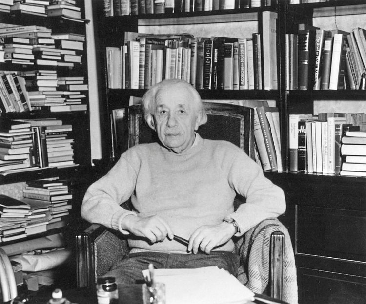 Ein alter Albert Einstein in einem Sessel, umgeben von Bücherregalen.