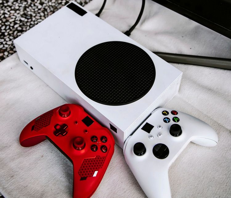 Eine weiße, rechteckige Spielekonsole liegt auf einer hellen Oberfläche. Daneben befinden sich zwei Controller: ein weißer und ein roter mit schwarzem Muster. Im Hintergrund sind Kabel sichtbar.