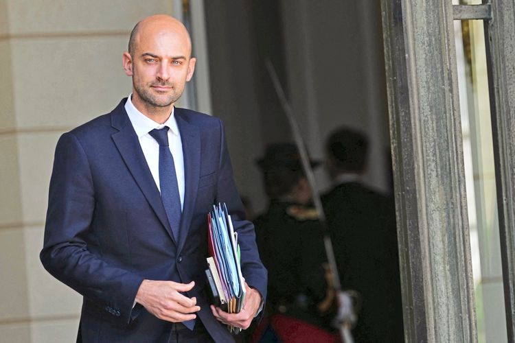 Ein Mann in einem dunkelblauen Anzug, der mehrere bunte Akten in der Hand hält, verlässt ein Gebäude mit einer großen Tür. Im Hintergrund sind unscharf Personen in Uniform zu erkennen.