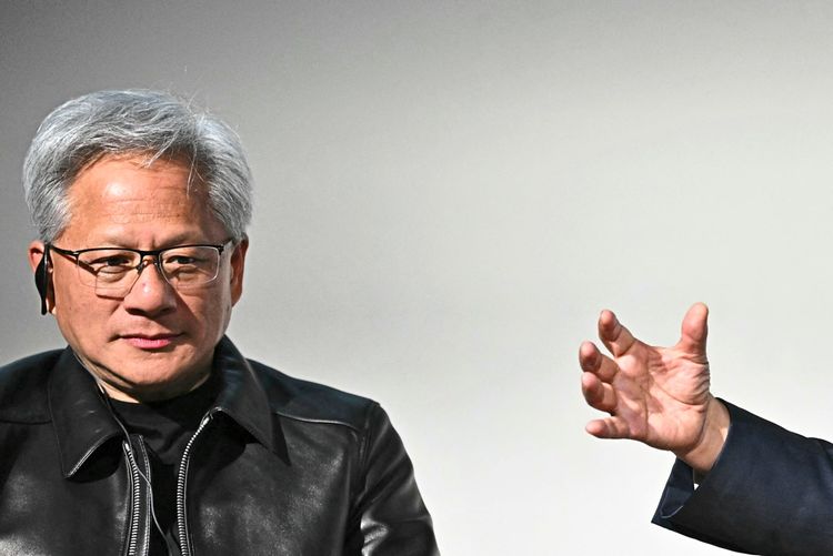 Ein Mann mit grauen Haaren und Brille, gekleidet in eine schwarze Lederjacke, sitzt vor einem neutralen Hintergrund. Rechts im Bild ist eine ausgestreckte Hand zu sehen, die auf ihn zeigt oder gestikuliert.