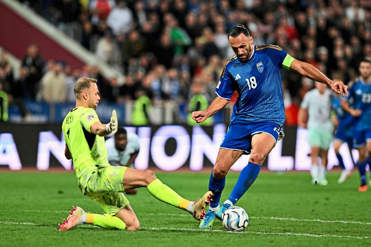 Zwei Fußballspieler kämpfen um den Ball während eines UEFA-Nations-League-Spiels zwischen Kosovo und Österreich in Pristina. Der Spieler im blauen Trikot trägt die Nummer 18, der Torwart im gelben Trikot die Nummer 01. Im Hintergrund sind Zuschauer zu sehen.