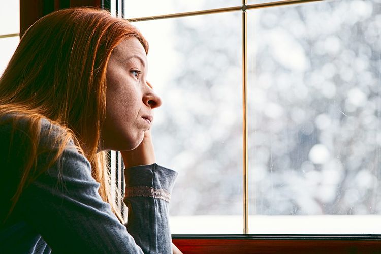 Frau blickt mit traurigem Gesichtsausdruck durch Fenster in Winterlandschaft