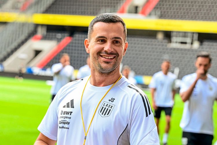 Ein Trainer des LASK Linz steht auf einem Fußballfeld in der Raiffeisenarena. Er trägt ein weißes Trainingsshirt mit Logos von Sponsoren und dem Vereinswappen sowie eine gelbe Pfeife um den Hals. Im Hintergrund sind unscharf Spieler und Tribünen erkennbar.