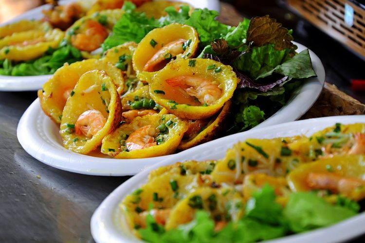 Vietnamesische gelbe Pfannkuchen in Kreisform auf Tellern, gefüllt mit Garnelen, Sojasprossen und Frühlingszwiebeln, serviert mit grünem Salat. Streetfood auf einem kulinarischen Markt in Ho-Chi-Minh-Stadt, Vietnam.