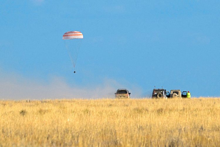 Kapsel an rot-weiß gestreiftem Fallschirm fällt über einem Getreidefeld Richtung Erdboden, zu sehen sind Autos der Rettungscrew.
