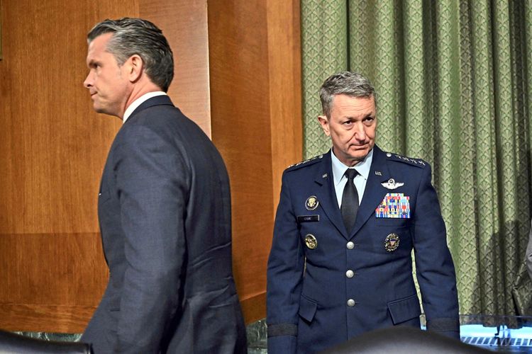 Zwei Männer verlassen einen Raum. Einer trägt einen dunkelblauen Anzug, der andere trägt eine Militäruniform mit Auszeichnungen und Abzeichen. Im Hintergrund sind grüne Vorhänge zu sehen.