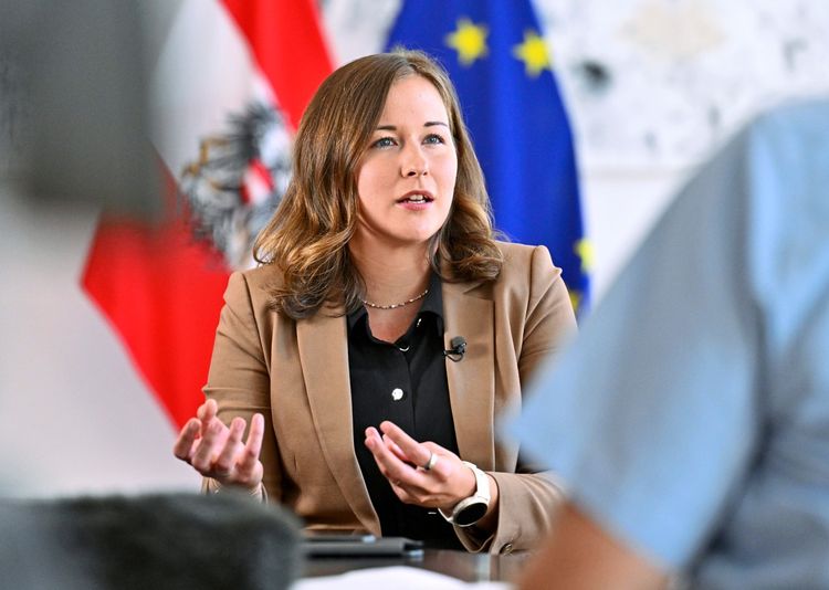 Eine Frau in braunem Blazer und schwarzer Bluse, vermutlich bei einem Interview, gestikuliert mit den Händen. Im Hintergrund sind unscharf die Flaggen von Österreich und der Europäischen Union zu sehen.