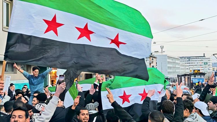 Jubelnde Menschenmenge in Kassel mit mehreren syrischen Oppositionsflaggen (grün-weiß-schwarz mit drei roten Sternen). Menschen feiern auf der Straße nach dem Sturz des Assad-Regimes. Gebäude und Straßenbahn im Hintergrund.