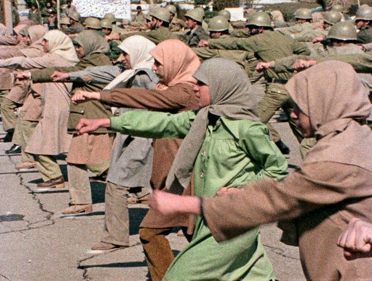 Frauen und Männer in militärischer Formation, mit ausgestreckten Armen in einer Übung. Die Frauen tragen Kopftücher und lange Mäntel, während die Männer Uniformen und Helme tragen. Die Szene wirkt wie ein Training oder eine Demonstration.