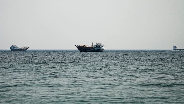 Drei Schiffe fahren auf ruhigem Wasser in der Straße von Hormus, aufgenommen vom Ufer in Musandam, Oman, bei leicht bewölktem Himmel.