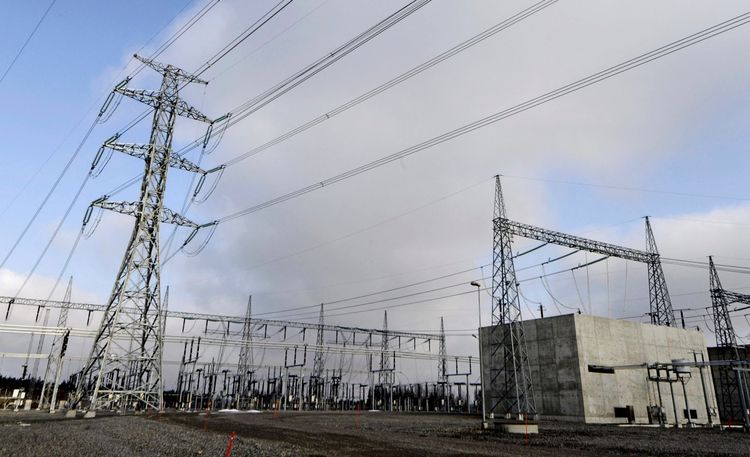Ein Transformatorstation mit Hochspannungsleitungen und metallischen Übertragungsstrukturen, umgeben von Kies. Der Himmel ist bewölkt.