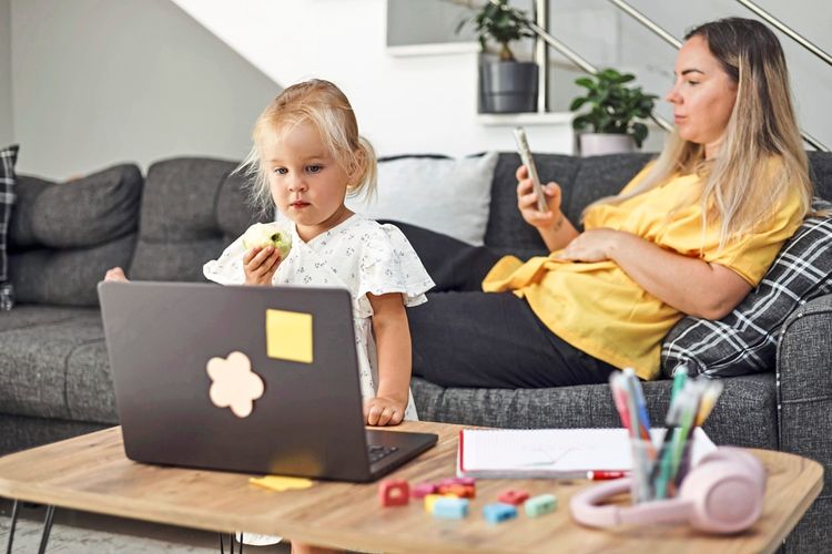 Ein kleines blondes Mädchen isst einen Apfel und schaut auf einen Laptop auf einem Tisch. Im Hintergrund sitzt eine Frau in einem gelben Oberteil, sieht auf ihr Smartphone und lehnt sich auf einer Couch zurück. Auf dem Tisch befinden sich bunte Spielzeuge, Kopfhörer und ein Notizbuch. Haustümliche Umgebung.