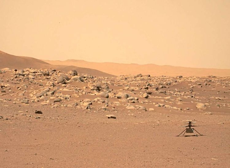 Ein sanfter Hügel auf dem Mars, darauf verstreute, runde Felsen. Rechts im Vordergrund der Marshelikopter.