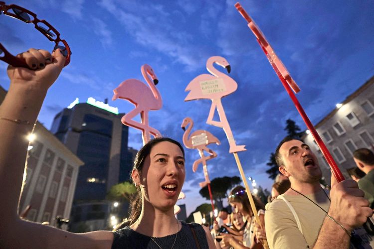 Protestierende halten rosafarbene Flamingo-Schilder und versammeln sich am Abend vor dem Büro des albanischen Premierministers in Tirana, um gegen den Bau von Luxushotels in geschützten Gebieten zu protestieren. Im Hintergrund sind Gebäude und der abendliche Himmel zu sehen.