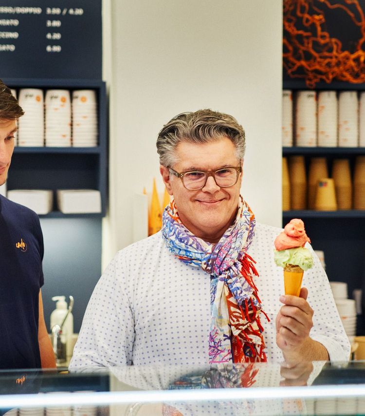 Carlo Maghakian hat graublondes Haar, trägt eine hellbraune Brille, einen bunten Schal und ein weißes Hemd mit grauen Punkten. In seiner linken Hand hält er ein Stanizel mit zwei Kugeln Eis.