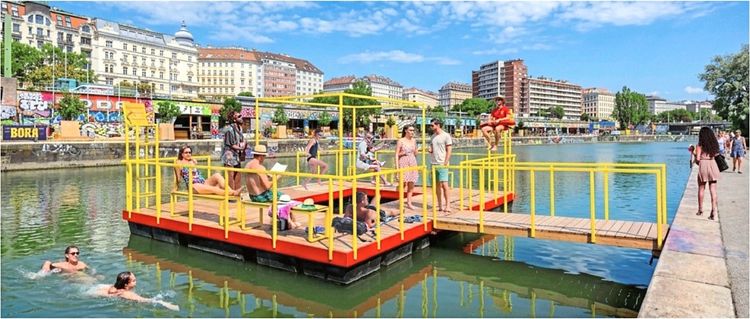 Menschen genießen einen sonnigen Tag an einem gelben, schwimmenden Ponton auf einem Fluss. Einige stehen und unterhalten sich, andere sitzen oder schwimmen im Wasser. Im Hintergrund sind Gebäude, Graffiti und Bäume zu sehen.