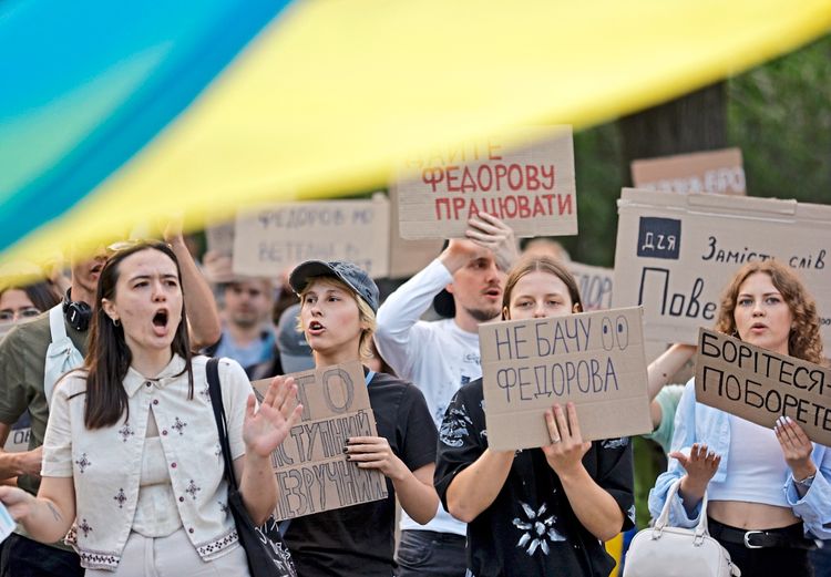 Eine Gruppe von Menschen protestiert in der Innenstadt von Kiew, Ukraine, mit Schildern, auf denen Botschaften wie „Fedorov ist der Verteidigungsminister“ und „Hände weg von Fedorov“ zu lesen sind. Sie fordern die Wiedereinsetzung von Mykhailo Fedorov als Verteidigungsminister. Im Vordergrund ist ein unscharfer Teil einer ukrainischen Flagge zu sehen.