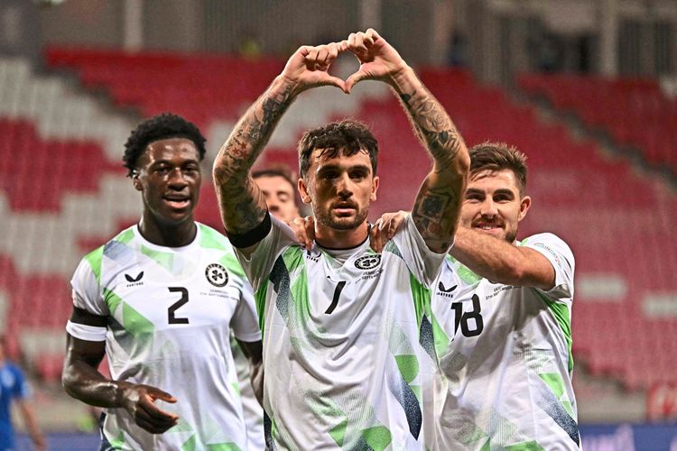 Drei Fußballspieler der irischen Nationalmannschaft während eines Spiels. Spieler in der Mitte (Rückennummer 7) zeigt eine Herzgeste mit den Händen, flankiert von zwei jubelnden Teamkollegen. Sie tragen weiße Trikots mit grünen Mustern. Im Hintergrund ein unscharfer Stadionbereich.