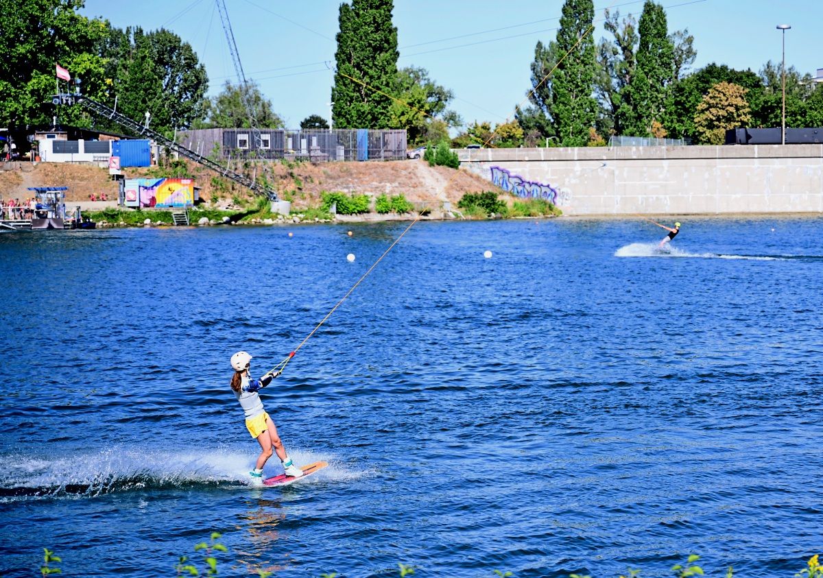 Eine Person fährt Wasserski auf einem See, gezogen von einem Seil eines Wasserskilifts. Im Hintergrund sind weitere Gebäude, Bäume und eine zweite Wasserskiläuferin zu sehen.