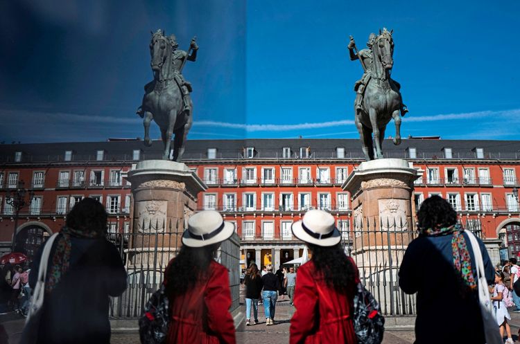 Personen spazieren über die Plaza Mayor in Madrid, Spanien, vorbei an Reiterstatuen und historischen Gebäuden unter einem klaren blauen Himmel.