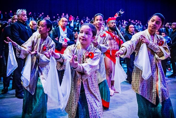 Vier Tänzerinnen in traditionellen Kleidern und mit weißen Tüchern in den Händen führen eine Choreografie auf, umgeben von Zuschauern in einem festlichen Veranstaltungsraum. Im Hintergrund sind weitere Personen in traditioneller Kleidung zu sehen. Die Szene findet während einer Feier zum 75. Jahrestag der Republik der Südmolukken in Den Bosch, Niederlande, statt.
