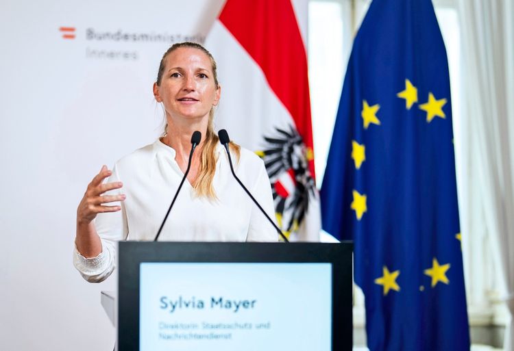 Eine Frau steht während einer Pressekonferenz am Rednerpult und gestikuliert. Im Hintergrund sind die Flagge Österreichs und die Europaflagge sichtbar. Auf dem Rednerpult steht der Name 