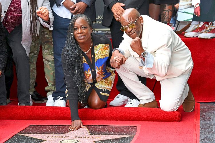 Zwei Personen knien auf einem roten Teppich neben Tupac Shakurs posthumer Stern auf dem Hollywood Walk of Fame. Die Frau trägt ein bunt gemustertes Kleid, der Mann ein weißes Outfit und eine Sonnenbrille. Weitere Menschen stehen im Hintergrund.