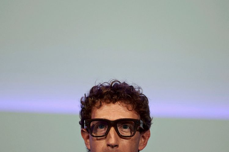 Ein Mann mit lockigem Haar trägt eine modern gestaltete Brille mit integriertem Display und spricht vor einem hellen Hintergrund.