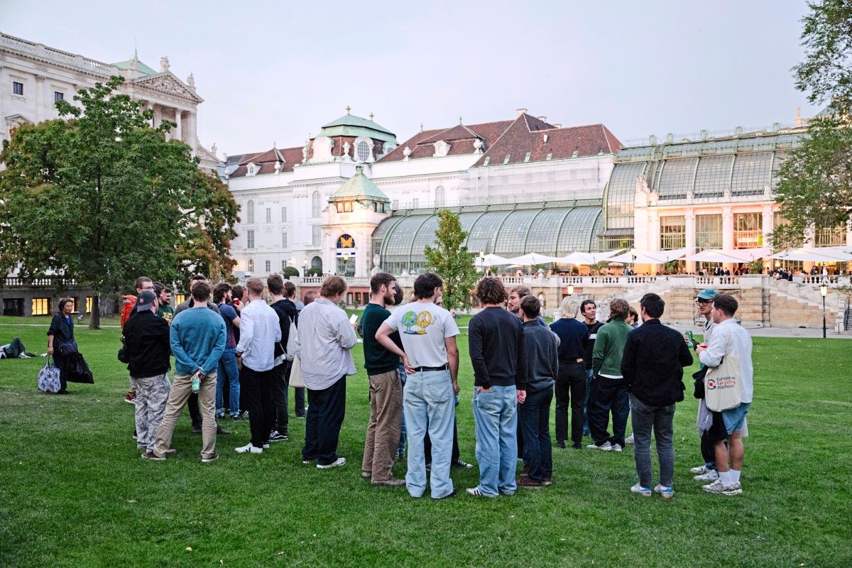 Eine Gruppe von Menschen steht auf einer Wiese vor einem historischen Gebäude mit Glasfassade und Terrasse. Es ist Abend, und die Szenerie wirkt entspannt und gesellig.