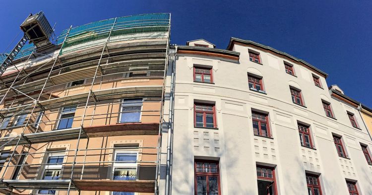 Ein mehrstöckiges Gebäude, dessen linke Seite von Baugerüsten umgeben ist, während die rechte Seite frisch renoviert und weiß gestrichen ist. Der Himmel ist klar und blau.