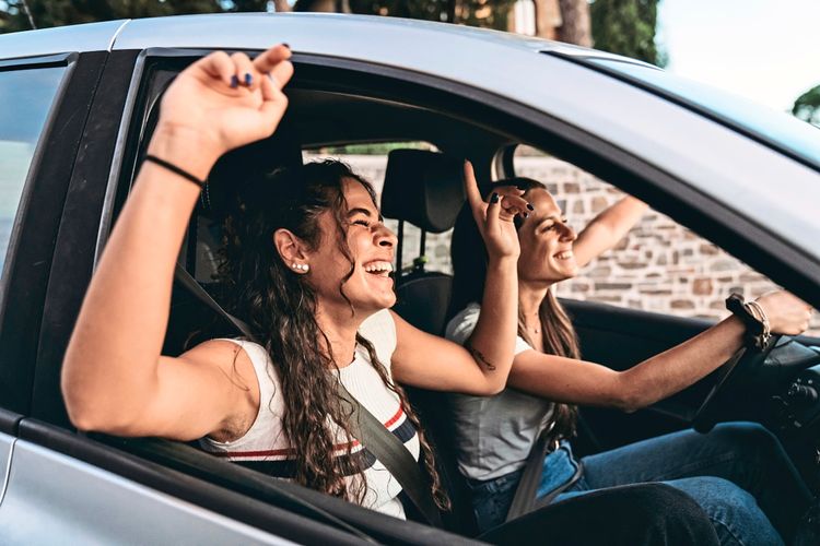 Zwei junge Frauen sitzen in einem Auto und heben fröhlich die Arme, während sie singen und gemeinsam Spaß haben.