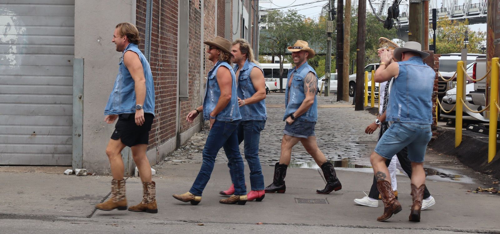 Sechs Männer in Jeans-Westen, mit Cowboy-Stiefeln und -Hüten auf einer Straße.