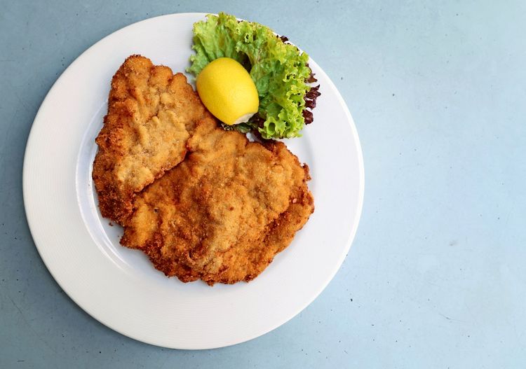Ein Wiener Schnitzel auf einem weißen Teller, serviert mit einem Stück Zitrone und grünem Salatgarnitur, auf einem hellblauen Tisch.