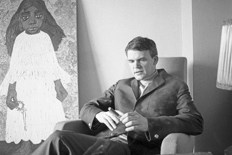 Ein Bild des Schriftstellers Milan Kundera mit einer Zigarre in der Hand vor einem Gemälde.