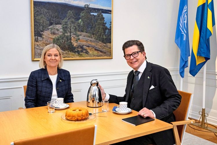 Zwei Personen sitzen an einem Tisch und führen ein Gespräch. Auf dem Tisch stehen Tassen, eine Kaffeekanne, Gläser und ein Kuchen. Im Hintergrund sind eine Landschaftsmalerei sowie die UN- und schwedische Flaggen zu sehen.