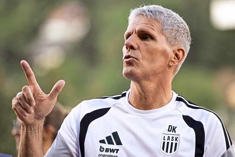 Ein Mann in einem LASK-Linz-Trainingsshirt, vermutlich der Trainer, gestikuliert und spricht während eines Fußballspiels. Hintergrund unscharf mit natürlicher Umgebung.