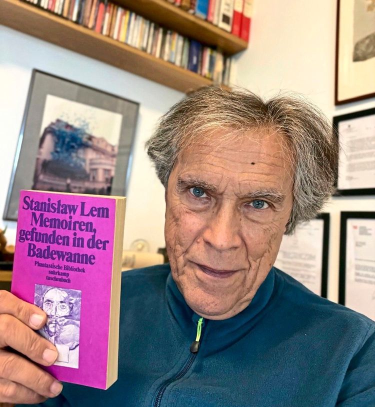 Ein Mann mittleren Alters hält das Buch „Stanislaw Lem – Memoiren, gefunden in der Badewanne“ mit violettem Einband in der Hand. Im Hintergrund sind Bücherregale und an der Wand hängende Bilder zu sehen.