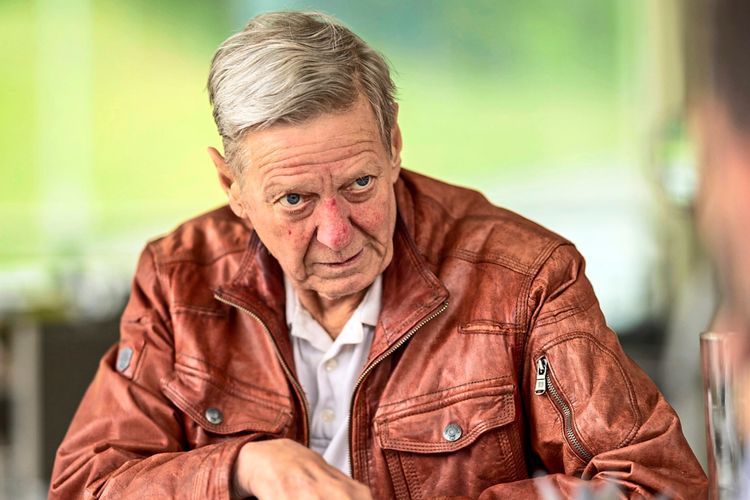 Ein älterer Mann mit grauen Haaren trägt eine braune Lederjacke und sitzt an einem Tisch. Er gestikuliert mit der Hand, während er mit einer Person außerhalb des Bildes spricht. Der Hintergrund ist unscharf und grün.